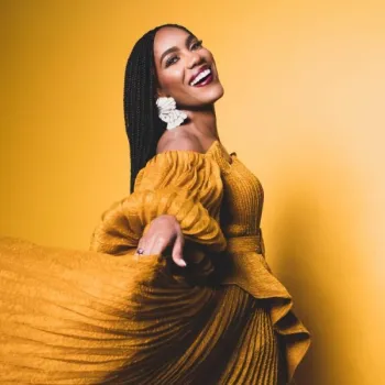 Woman in a textured, mustard yellow off-the-shoulder dress poses against a matching yellow background, smiling widely and holding out part of her dress.