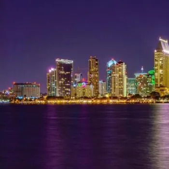Nighttime view of the San Diego skyline with illuminated buildings reflecting on the water, and a lit bridge visible on the left.