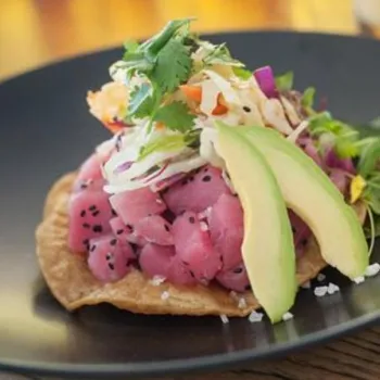 A tostada topped with diced raw tuna, sliced avocado, shredded vegetables, sesame seeds, and fresh herbs, served on a black plate.