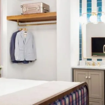 A neatly made bed with a plaid-patterned pillow, an open closet with clothes and suitcase, a small vanity area, and a desk with a lamp in a modern hotel room.