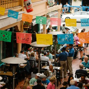 Outdoor restaurant scene with people dining and socializing under colorful papel picado banners on a busy street.