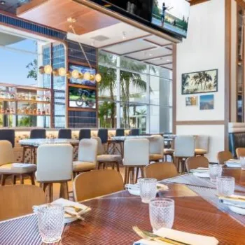 A modern restaurant interior with neatly set tables, upholstered chairs, a bar area, and large windows letting in natural light.