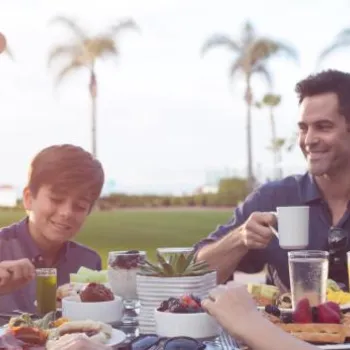 A family of four sits at an outdoor table enjoying breakfast with fruit, juice, and coffee, with palm trees and greenery in the background.