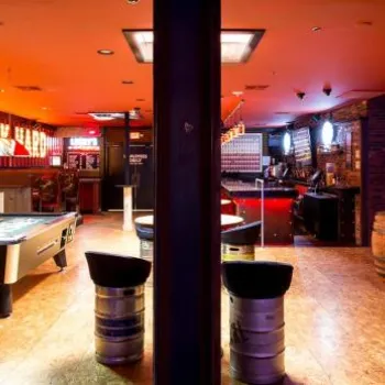 A bar with neon signs, pool table, barrels, and chain-link dividers; wood flooring and a warm, orange ambient lighting.
