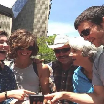 Five people stand outdoors in sunlight, smiling and laughing as they look at a device held by one person. Trees and tall buildings are visible in the background.
