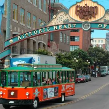 A green and orange trolley drives under the "Historic Heart of San Diego" arch in the Gaslamp Quarter, surrounded by city buildings and shops.