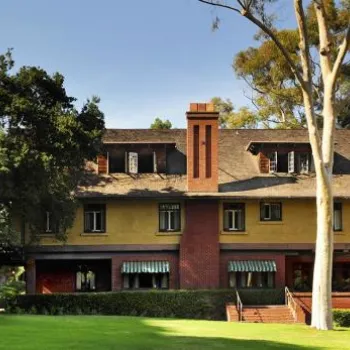 Large two-story house with brick and yellow exterior, surrounded by trees and green lawn under a blue sky.