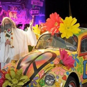 Two people in hippie costumes stand by a colorful, flower-covered VW Beetle with peace signs, in a room decorated with 1960s-themed artwork and signs.