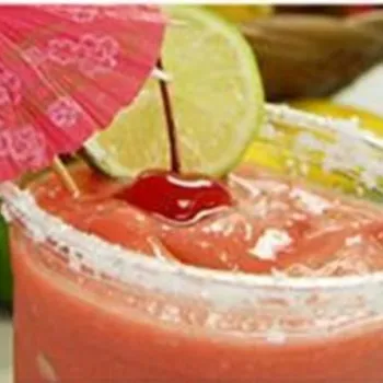 A frozen pink drink in a plastic cup with a salted rim, garnished with a lime slice, cherry, and a pink paper umbrella. Fruits are visible in the background.