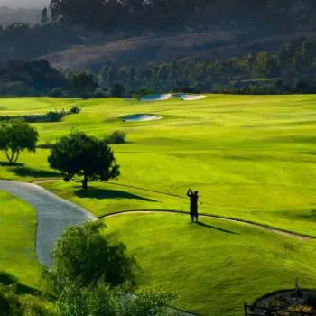 A golf course with green fairways, sand bunkers, scattered trees, and a winding path set against a backdrop of hills and forest.