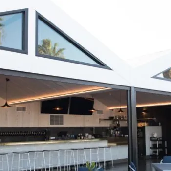 Modern open-air bar with a long counter, barstools, and minimalist decor; triangular roof windows and outdoor seating visible.