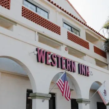 Exterior view of a white building with arches labeled "Western Inn," displaying the American flag and other flags, with palm trees nearby.