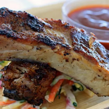 Two grilled pork ribs served on a bed of coleslaw with a small cup of barbecue sauce on the side.