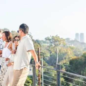 A group of people stand and converse on an outdoor balcony with a city skyline and greenery visible in the background.