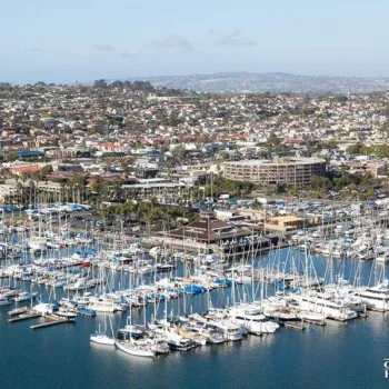 Aerial view of a marina filled with docked sailboats and yachts, with a coastal city and hills in the background under a partly cloudy sky.