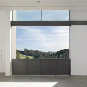 A modern white-walled room with large windows overlooks a scenic landscape with hills and trees under a blue sky; minimal art hangs on the walls.