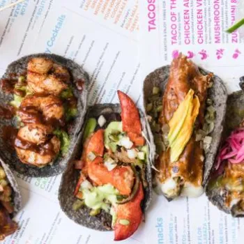 Five assorted tacos with various toppings are arranged in a row on a colorful menu, accompanied by drinks with lime garnishes.