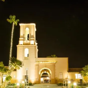 A historic building with a central tower and palm trees wrapped in string lights, illuminated at night with a tall modern building in the background.