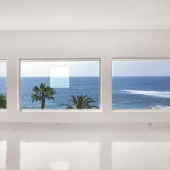 An empty, white room with large windows offering a panoramic ocean view, palm trees, and clear blue sky outside.