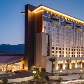 A modern high-rise hotel with illuminated windows and exterior lights, set against a mountainous backdrop at dusk.
