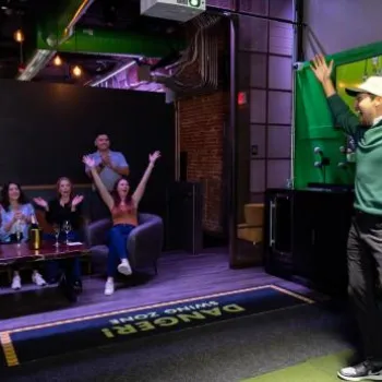 A man celebrates a golf swing in an indoor golf simulator while a group of people cheers from a nearby couch in a sports-themed lounge.