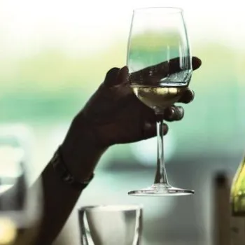 A person holds a glass of white wine at a table set with more glasses and a wine bottle, with a blurred background.