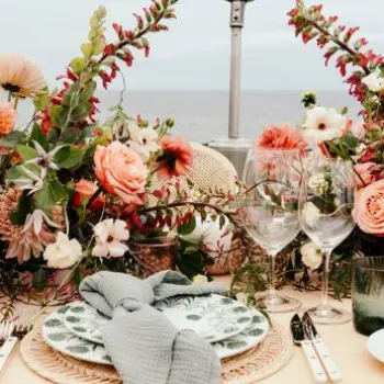 A formal outdoor table setting with floral centerpieces, glassware, plates, and cutlery arranged neatly against a seaside background.