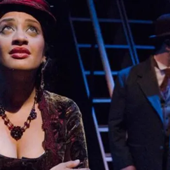A woman in vintage clothing and a hat looks upward with a serious expression, while a man in a suit and hat stands behind her on a dimly lit stage.