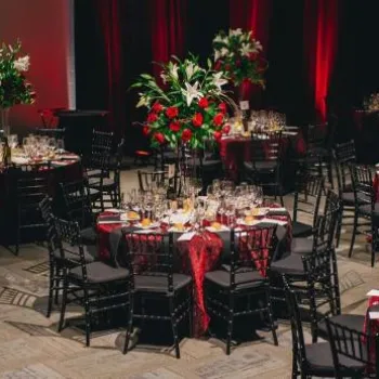 Ballroom set for an event with round tables covered in red cloths, each adorned with tall floral centerpieces and surrounded by black chairs.