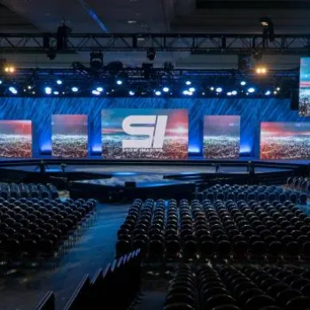 Large conference hall with rows of empty chairs facing a stage, multiple large screens displaying the same logo, and blue stage lighting.