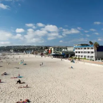 People relax and walk on a sandy beach near the ocean, with hotels and buildings lining the shoreline under a partly cloudy sky.