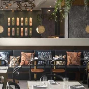Modern restaurant interior with black chairs, wooden tables, patterned cushions, hanging plants, and decorative wall panels, with natural light coming through large windows.