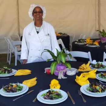 A chef in a white uniform and hat stands smiling in front of round tables set with salads, yellow napkins, and floral centerpieces under a white tent.