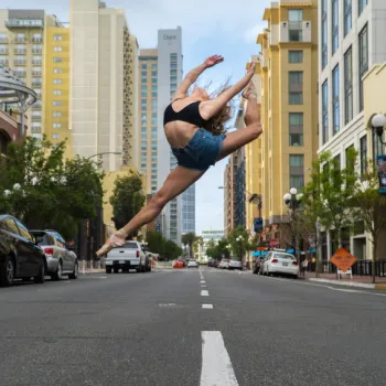 A dancer performs a high leap in the middle of an empty city street, surrounded by tall buildings and parked cars.