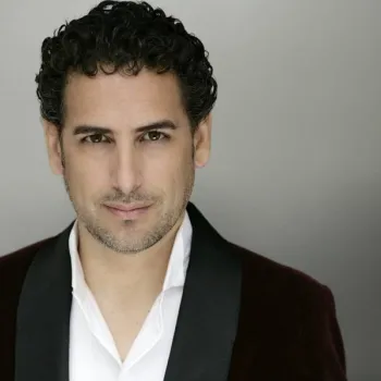 A man with short curly dark hair and light facial stubble wears a white shirt and a dark blazer, facing the camera against a plain gray background.
