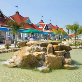 Outdoor shopping area with red-roofed buildings, a central stone fountain, clear water, and people walking around on a sunny day.