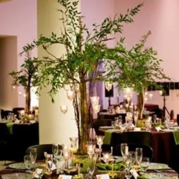 Round tables set for an event with glassware and plates, each table decorated with tall centerpieces of greenery and hanging lights in a spacious indoor venue.