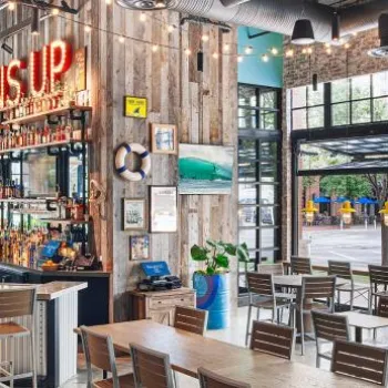 Bright, modern restaurant with nautical decor, a central bar, wooden tables and chairs, large windows, and surfboards on the wall.