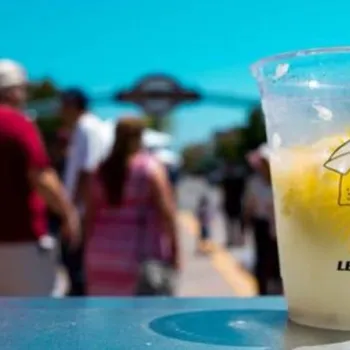 A nearly empty plastic cup of lemonade with a straw sits on a ledge, with a crowd of people walking outdoors in the background.