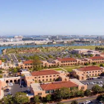 Aerial view of a campus with red-roofed buildings, parking lots, greenery, and water in the background, with a city skyline and airport visible in the distance.