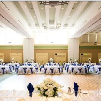 Large banquet hall set up for an event, with round tables covered in white tablecloths and blue chair sashes, floral centerpieces, and bright overhead lighting.