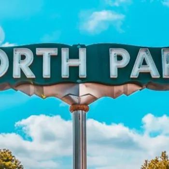 A large retro-style sign reading "NORTH PARK" stands against a blue sky with some clouds, trees, and colorful buildings in the background.