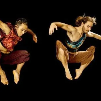 Two dancers wearing colorful tops and loose pants are captured mid-air against a black background, with arms bent and knees raised.