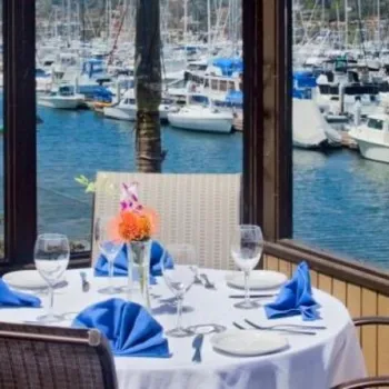 A waterfront restaurant table is set with blue napkins, glassware, and a flower centerpiece, overlooking a marina with numerous docked boats.