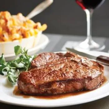 A grilled steak on a white plate with greens, a serving of macaroni and cheese in a bowl, and a glass of red wine in the background.