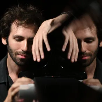 A man with closed eyes rests his head and hands on a reflective black surface, creating a mirrored image of his face and hands.