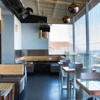 Modern restaurant interior with wooden tables and chairs, large windows offering an ocean view, and natural light filling the space.