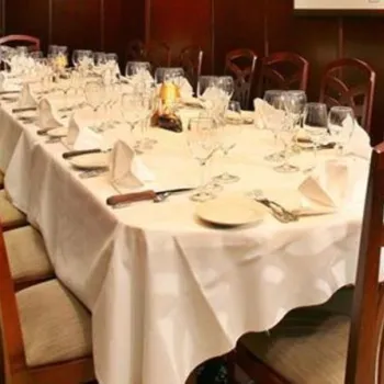 A long dining table set for a formal meal with white tablecloth, plates, glassware, and cutlery, surrounded by wooden chairs in an elegant room.