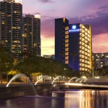 A cityscape at dusk features illuminated Wyndham hotels, high-rise buildings, trees, and fountains near a water feature under a purple and orange sky.