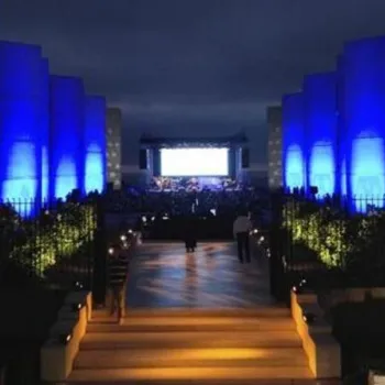 Outdoor concert at night with blue-lit concrete buildings flanking a central walkway leading to a stage with a large screen in the background.